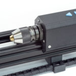 Sub spindle on bench top coil winder. The sub spindle has a keyless 3 jaw chuck and two blue buttons on top to move it left and right for mandrel tensioning.