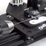 A close up of a coil winder showing a thin stainless wire being wound over a mandrel under tension. A keyless three jaw chuck is holding the left side of the mandrel.